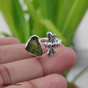 May include: A silver ring with an olive-green gemstone and a bee motif. The gemstone is set in a bezel, and the bee is intricately detailed. The ring's design is open, with the bee on one side.
