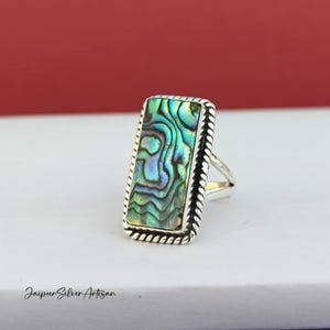 May include: A silver ring featuring a rectangular abalone shell inlay. The shell displays a swirling pattern of green, blue, and purple.