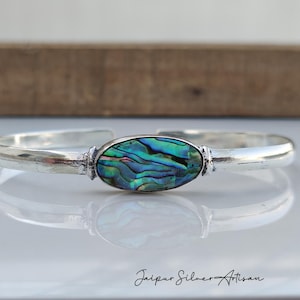 May include: A silver cuff bracelet featuring an oval abalone shell centerpiece with iridescent blues, greens, and purples. The bracelet has a polished finish and is displayed on a reflective surface. The Jaipur Silver Artisan logo is visible.