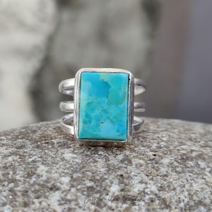 May include: A silver ring with a rectangular turquoise stone. The turquoise is a vibrant blue-green color. The ring has a triple-band design. The text "Jaipur Silver Artisian" is at the bottom.