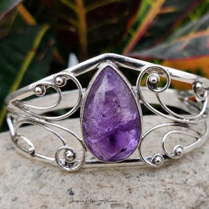 May include: A silver cuff bracelet featuring a central pear-shaped purple amethyst gemstone. The metal band is crafted with openwork scroll patterns and small silver spheres. The text Jaipur Silver Artisan is printed at the bottom of the image.
