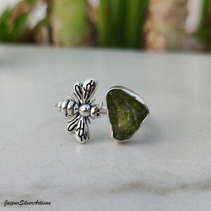 May include: A silver ring with a bee motif and a large, irregularly shaped, pale green gemstone. The ring is displayed against a neutral backdrop, highlighting the intricate details of the jewellery. The ring is a unique piece of jewellery.