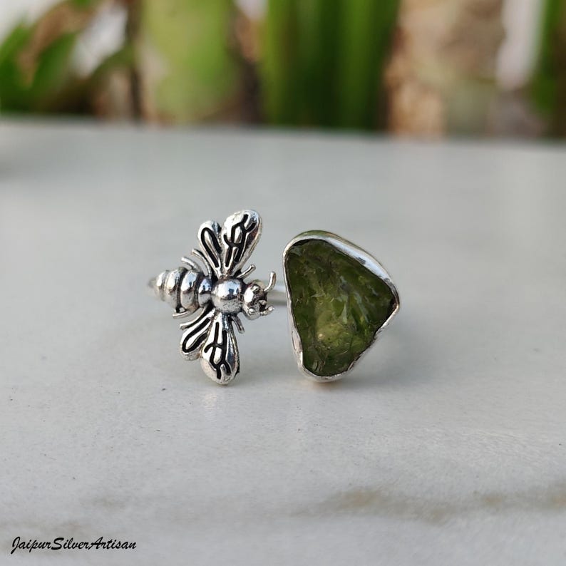 May include: A silver ring with a bee motif and a large, irregularly shaped, pale green gemstone. The ring is displayed against a neutral backdrop, highlighting the intricate details of the jewellery. The ring is a unique piece of jewellery.