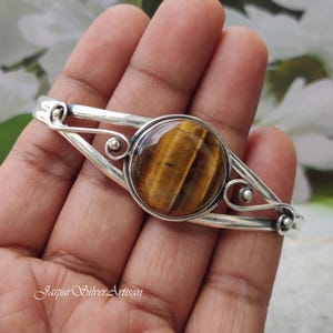May include: A silver bangle bracelet with a large, round, brown tiger's eye gemstone set in the center. The bracelet has a delicate, openwork design.