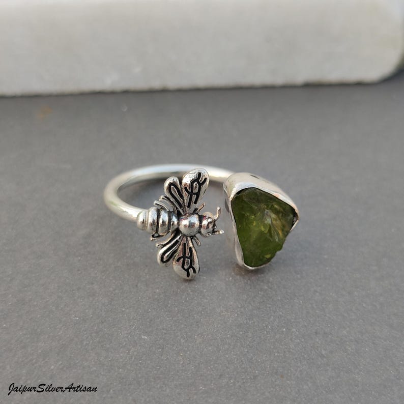 May include: A silver ring with a bee motif and a raw, olive-green peridot gemstone. The ring has an open design, with the bee on one end and the gemstone on the other. The ring is displayed against a neutral grey background.