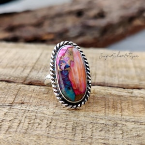 Op de afbeelding: Een zilveren ring met een grote ovale edelsteen. De edelsteen is een mix van roze, oranje, bruin en blauwe kleuren. De ring heeft een touwachtig ontwerp rond de rand van de zetting. "Jaipur Silver Artisan" staat op de achtergrond.