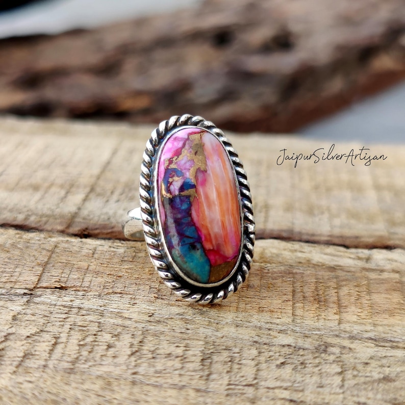 Op de afbeelding: Een zilveren ring met een grote ovale edelsteen. De edelsteen is een mix van roze, oranje, bruin en blauwe kleuren. De ring heeft een touwachtig ontwerp rond de rand van de zetting. "Jaipur Silver Artisan" staat op de achtergrond.
