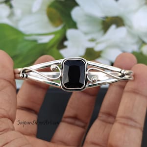 May include: A silver bangle bracelet with a black gemstone set in a square shape. The bracelet has a delicate, ornate design.