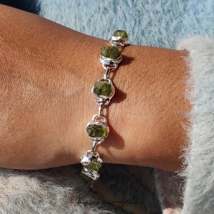 May include: A silver bracelet with five raw, olive-green peridot stones. Each stone is set in a silver bezel and linked by a silver chain. The bracelet is shown on a wrist, set against a soft blue background. The peridot stones are a vibrant green.