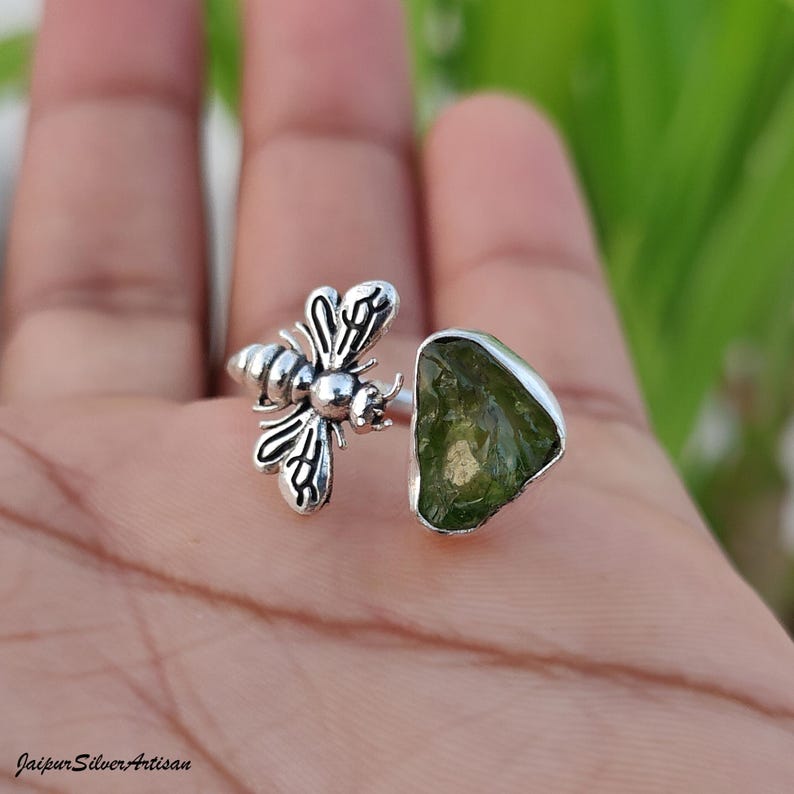 May include: A silver ring with a bee motif and a green gemstone. The bee has detailed wings and body segments. The gemstone is an irregular shape. The ring is on a hand, with the Jaipur Silver Artisan logo visible.