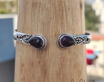 May include: A silver bangle bracelet with two dark purple gemstone accents. The bracelet has a delicate, ornate design.