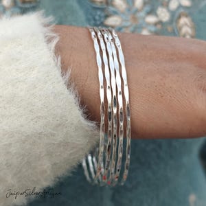 May include: A set of five silver bangles with a hammered texture. The bangles are stacked on a person's wrist.