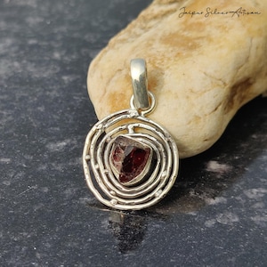 May include: A silver pendant with a raw, dark red gemstone at its center. The gemstone is set within a circular, textured silver frame. The pendant has a loop at the top for a chain. The text "Jaipur Silver Artisan" is in the upper right corner.