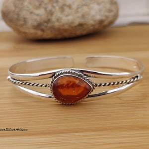 May include: A silver cuff bracelet with a teardrop-shaped amber stone. The amber is a warm orange color, set in a silver bezel with a twisted rope design. The bracelet is open-ended and rests on a wooden surface.