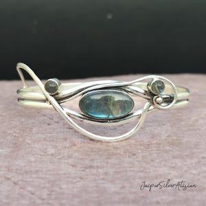 May include: A silver bracelet with a blue labradorite gemstone and two smaller silver gemstones. The bracelet has a delicate, flowing design.