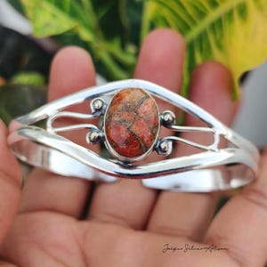 Può includere: Un bracciale a fascia in argento lucido con una pietra ovale dai motivi arancioni e marroni incastonata al centro. La fascia metallica presenta un design diviso con sfere d'argento decorative che fiancheggiano l'incastonatura. Il testo Jaipur Silver Artisan è visibile.