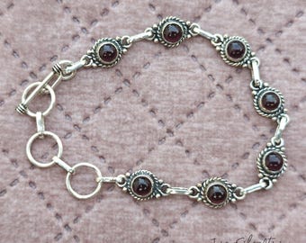 May include: A silver bracelet with a toggle clasp and seven dark red gemstone accents. The bracelet is made of linked silver circles and has a decorative design around each gemstone.