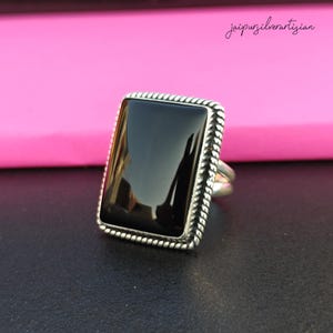 May include: A silver ring with a black rectangular gemstone set in a rope-like border.