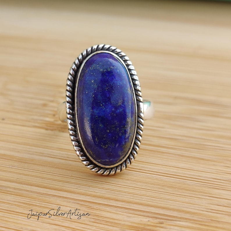 Op de afbeelding: Een zilveren ring met een grote ovale lapis lazuli-edelsteen. De ring heeft een touwachtig ontwerp rond de rand van de zetting.