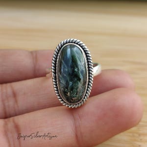 Op de afbeelding: Een zilveren ring met een ovale groene en zwarte edelsteen, gezet in een touwachtige rand.