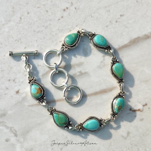 May include: A silver bracelet featuring turquoise teardrop-shaped stones. The bracelet has a toggle clasp and is connected by silver links. The turquoise stones have brown inclusions. The words "Jaipur Silver Artisan" are at the bottom of the image.