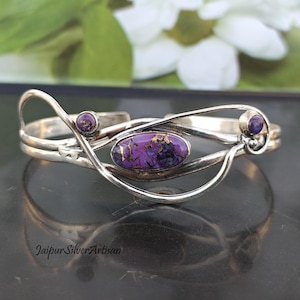 May include: A silver bracelet featuring a purple and gold gemstone set in a delicate, swirling design. The bracelet is open and adjustable.