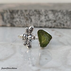 May include: A silver ring with a bee motif and a raw, light green peridot gemstone. The ring's bee has detailed wings and body. The peridot is set in a silver bezel. The ring is shown on a reflective surface. The ring is a unique piece of jewellery.
