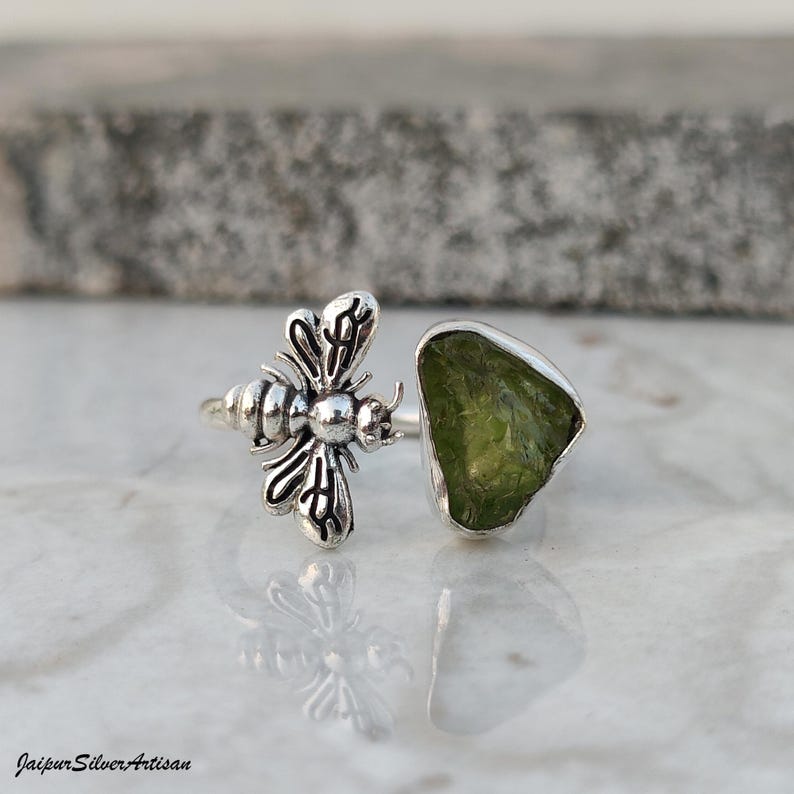 May include: A silver ring with a bee motif and a raw, light green peridot gemstone. The ring's bee has detailed wings and body. The peridot is set in a silver bezel. The ring is shown on a reflective surface. The ring is a unique piece of jewellery.