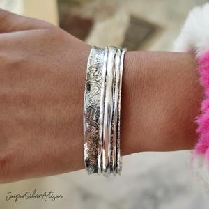 May include: A set of four silver bangles with an intricate floral design. The bangles are stacked on each other and are worn on a person's wrist.