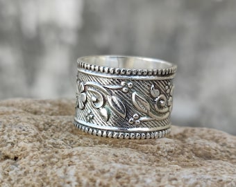 May include: A wide, silver ring with an intricate floral design. The ring features detailed carvings of flowers and leaves, with a textured background. The edges are lined with small, beaded accents. The ring is displayed on a rough, tan surface.