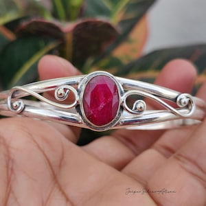May include: A silver cuff bracelet featuring a central oval-shaped faceted deep pink ruby gemstone. The metal band displays a polished finish with decorative swirling wire accents on both sides of the stone. The text Jaipur Silver Artisan is visible.