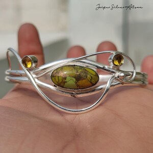 May include: A silver bracelet with an oval, green and brown stone in the center, flanked by two smaller yellow stones. The bracelet has a unique, flowing design.