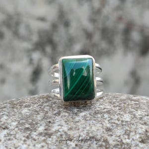 May include: A silver ring featuring a rectangular, vivid green malachite gemstone. The ring has a triple-band design. The ring is set against a neutral, textured background. A piece of jewellery.