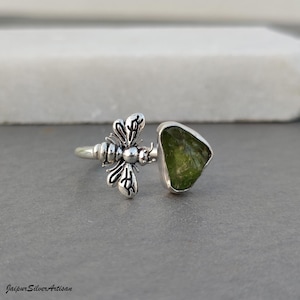 May include: A silver ring with a bee motif and a large, irregular-shaped, light green gemstone. The ring's band is thin, and the bee has detailed wings and body. The gemstone is set in a silver bezel. The ring is likely made of sterling silver.