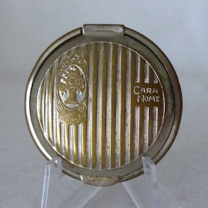 May include: A silver and gold compact mirror with a striped design and the words "Cara Nome" engraved on the side. The compact has a floral design with the words "Lucien Lelong New York" in the center.