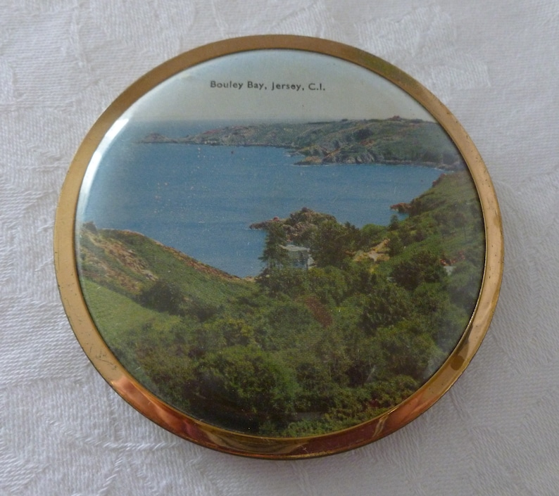 Puede incluir: Un espejo compacto redondo con un borde dorado, que presenta una imagen de postal vintage de Bouley Bay, Jersey, C.I. La imagen muestra una vista panor&aacute;mica de una bah&iacute;a con colinas verdes y agua azul.