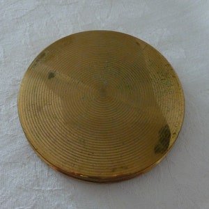 Puede incluir: Un compacto redondo de color dorado con un patr&oacute;n de c&iacute;rculos conc&eacute;ntricos en la tapa.