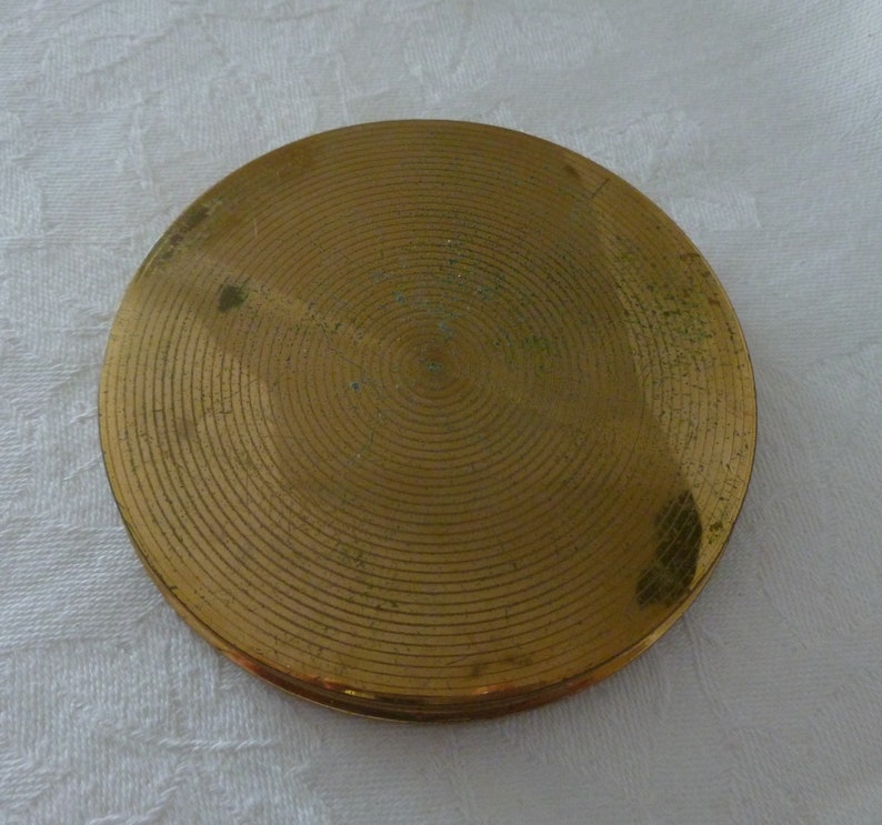 Puede incluir: Un compacto redondo de color dorado con un patr&oacute;n de c&iacute;rculos conc&eacute;ntricos en la tapa.