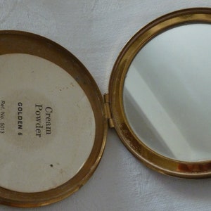 Puede incluir: Un estuche dorado con un espejo y una etiqueta blanca que dice "Cream Powder GOLDEN 6 Ref. No. 5013"