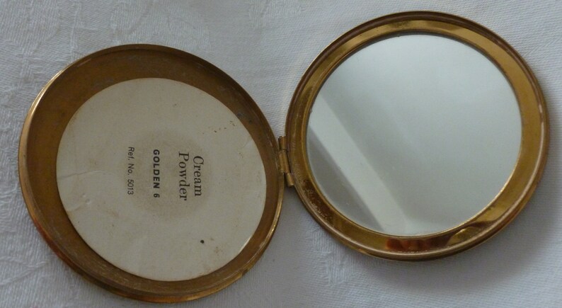 Puede incluir: Un estuche dorado con un espejo y una etiqueta blanca que dice "Cream Powder GOLDEN 6 Ref. No. 5013"