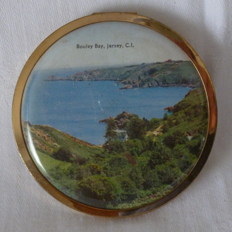 Puede incluir: Un espejo compacto de color dorado con una imagen redonda, blanca y azul de una bah&iacute;a con colinas verdes y el texto "Bouley Bay, Jersey, C.I."