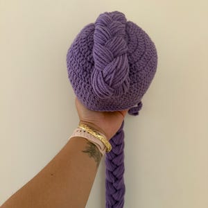 May include: A handmade, purple crocheted ball with a braided top and a long, braided tail. The ball is held in a hand against a plain white background. The crochet work is detailed and the color is a vibrant shade of purple.