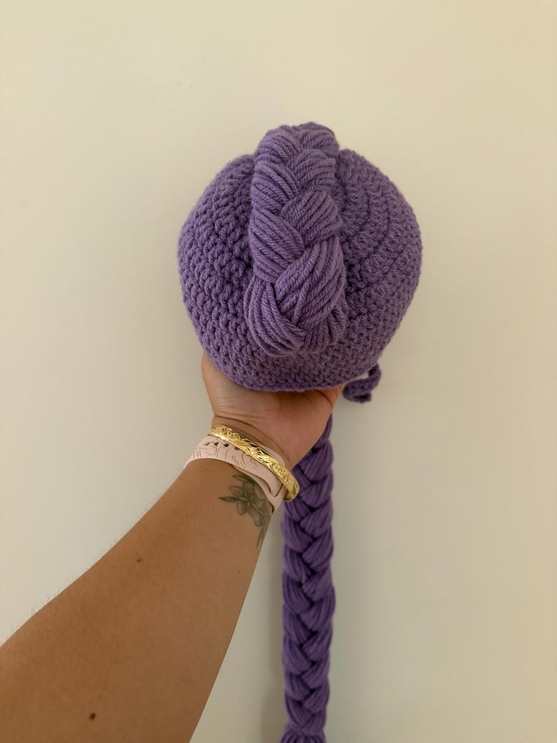 May include: A handmade, purple crocheted ball with a braided top and a long, braided tail. The ball is held in a hand against a plain white background. The crochet work is detailed and the color is a vibrant shade of purple.