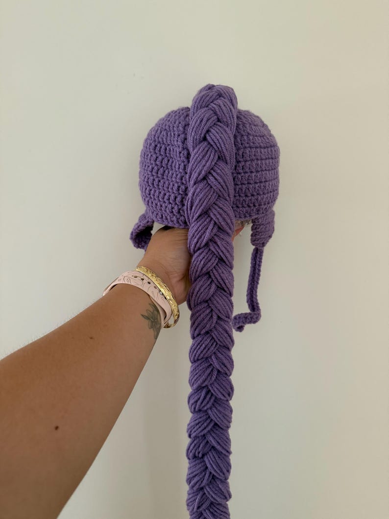 May include: A purple crocheted hat with a long, braided extension. The hat features ear flaps and a braided tail, all in a solid shade of purple. The hat is held up against a white background.