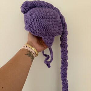 May include: A purple crocheted hat with a long, braided tail. The hat is a solid color and appears to be handmade. The braid is thick and hangs down. The hat is likely for a child or a costume.