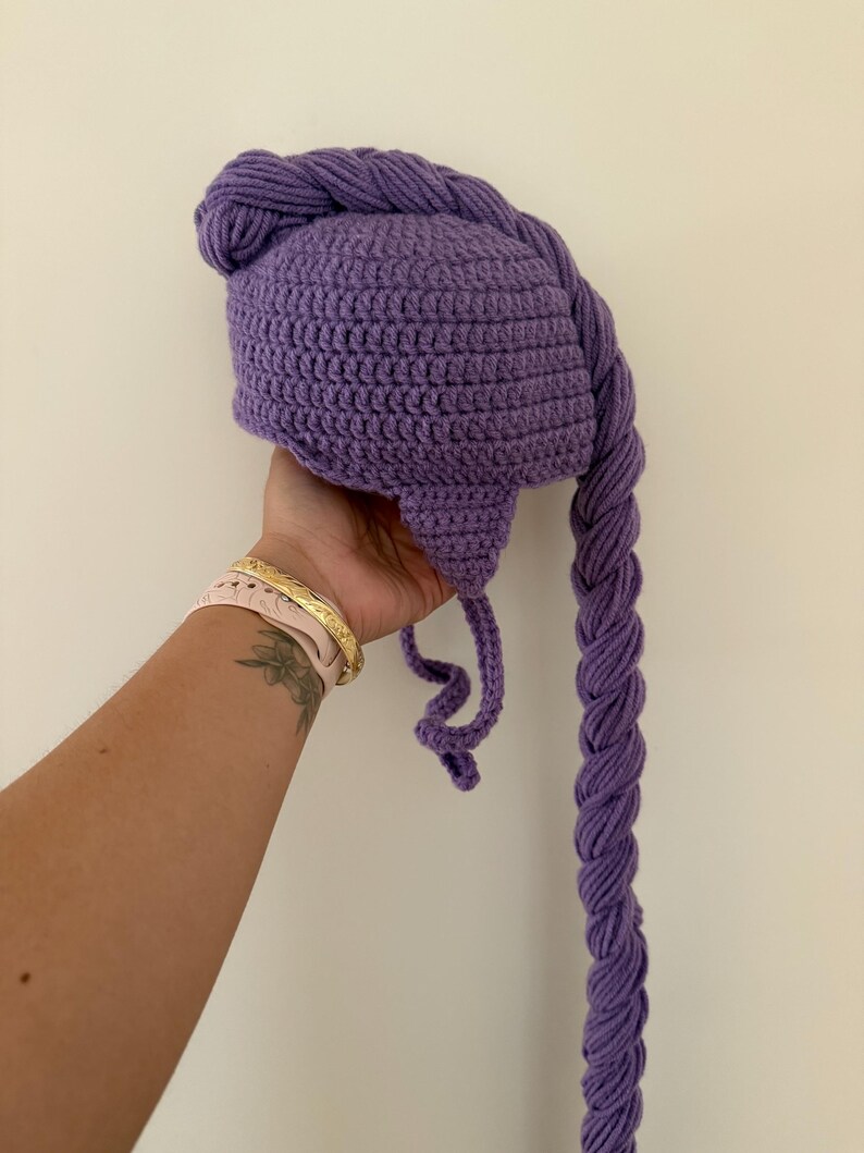 May include: A purple crocheted hat with a long, braided tail. The hat is a solid color and appears to be handmade. The braid is thick and hangs down. The hat is likely for a child or a costume.