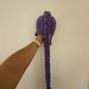 May include: A purple crochet hat with a long, braided tail. The hat is a solid color, and the braid is thick and detailed. The tail hangs down, ending in a fringe. The hat is held up against a white wall.