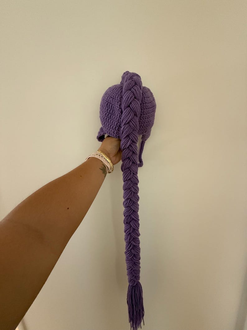 May include: A purple crochet hat with a long, braided tail. The hat is a solid color, and the braid is thick and detailed. The tail hangs down, ending in a fringe. The hat is held up against a white wall.