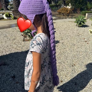 May include: A purple crocheted hat with a long, braided tail. The hat is a vibrant shade of lavender and features ear flaps. The braided tail hangs down the back. The hat is worn by a person.