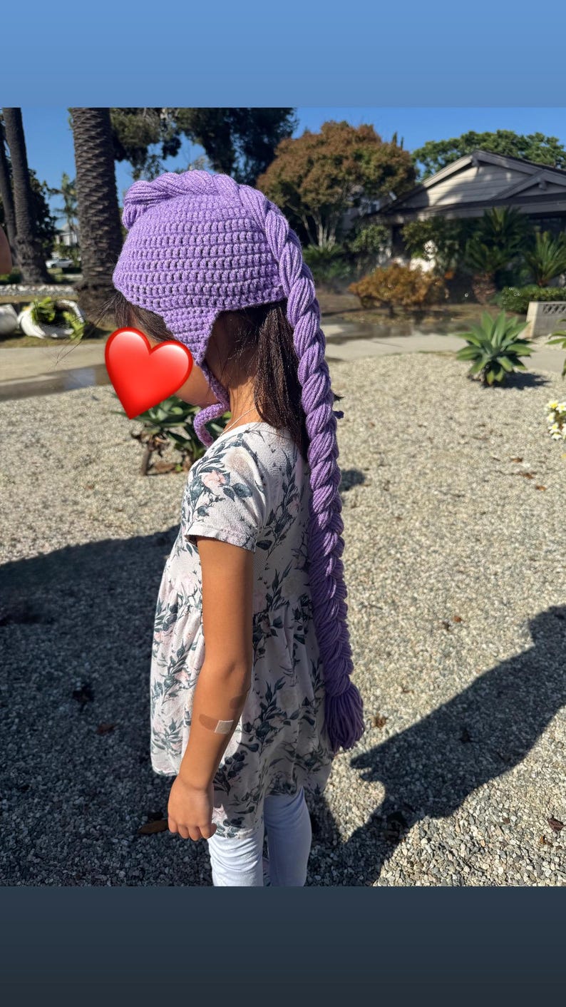 May include: A purple crocheted hat with a long, braided tail. The hat is a vibrant shade of lavender and features ear flaps. The braided tail hangs down the back. The hat is worn by a person.
